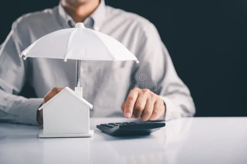 Man calculating finances beside a small house protected by an umbrella, symbolizing business insurance and financial protection. 