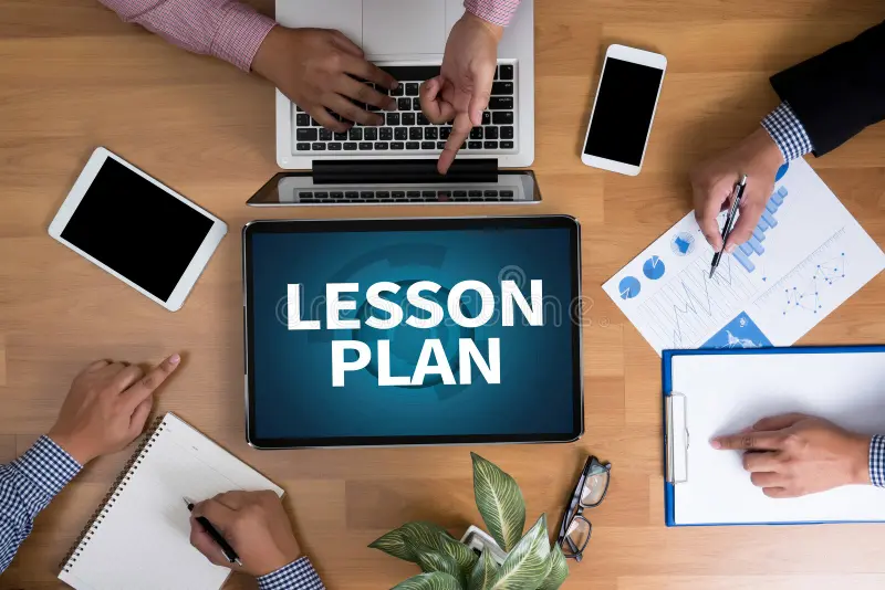 Lesson plan displayed on a tablet with a business team working on laptops, financial reports, charts, and notes around a table.