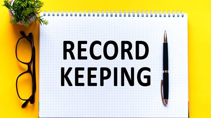 Recordkeeping concept featuring a notebook labeled โRecord Keepingโ with a pen, eyeglasses, and office supplies.