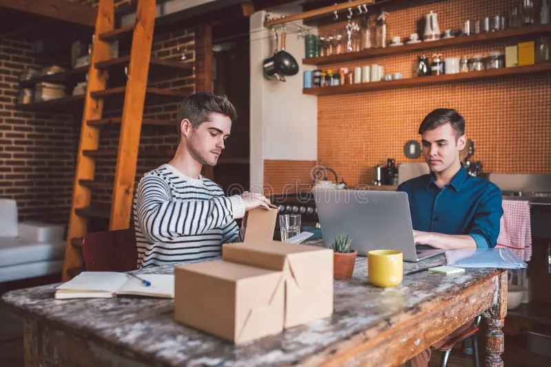 Two home-based business owners packing shipping boxes and working on a laptop at a kitchen table