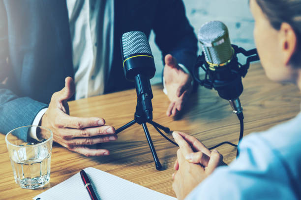Two people recording a podcast interview at a wooden table with studio microphones, a glass of water, and a notebook, representing how to start a podcast editing business